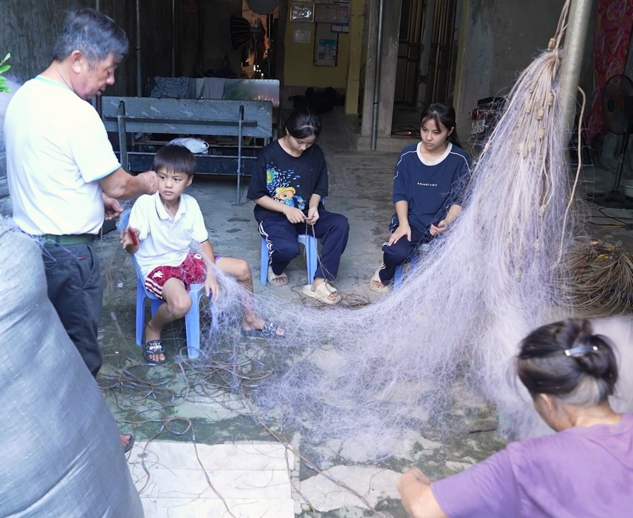 Ông Nguyễn Văn Cưng, bà Bùi Thị Tương cùng ba cháu nội miệt mài đan lưới thuê – công việc chính mang lại thu nhập cho cả nhà.